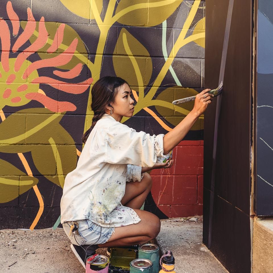 A woman in a paint-splattered shirt paints a mural on a wall with a floral background.