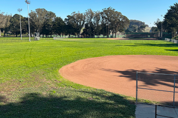 A sunny day at an empty baseball field surrounded by green grass and trees.