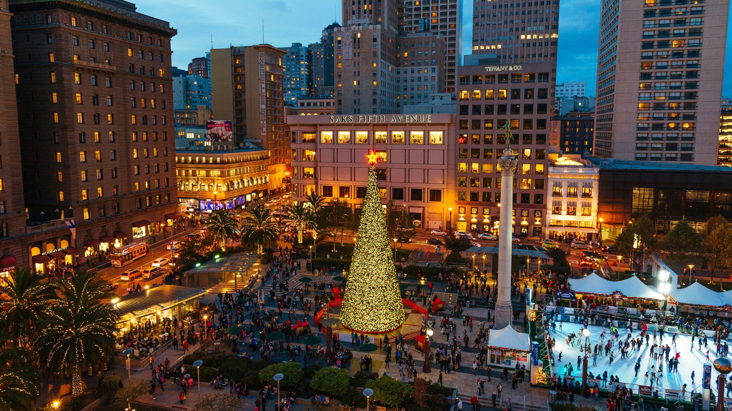 A large Christmas tree glows in a busy city square at dusk, surrounded by tall buildings and crowds of people.