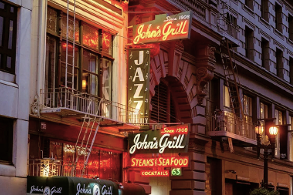 Neon signs for John's Grill and JAZZ glow on the exterior of a historic city building at dusk.