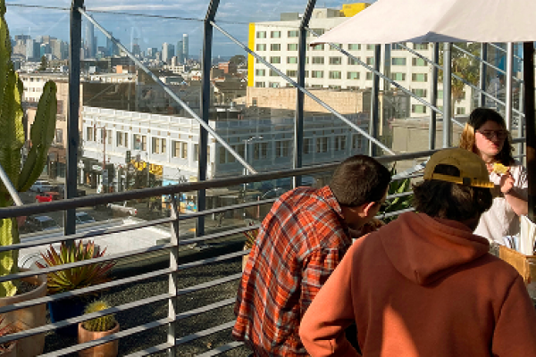 Three people converse on a sunny rooftop patio with city buildings and a large cactus in the background.
