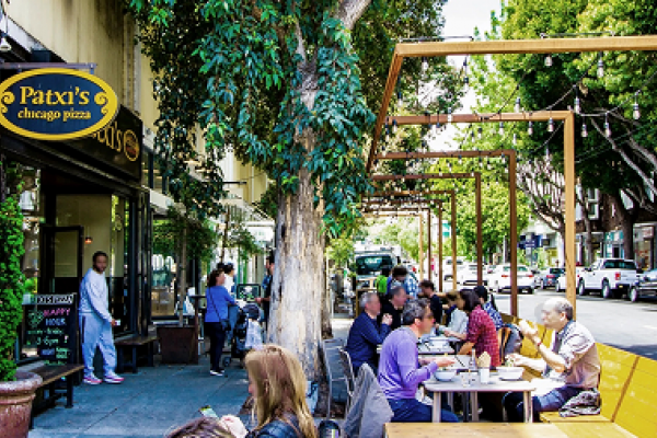 People dining outdoors at Patxi's Chicago Pizza on a sunny, tree-lined street with parked cars.