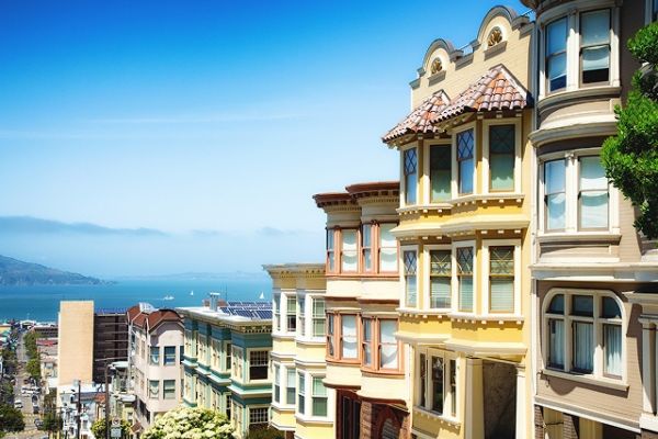Colorful, classic San Francisco houses on a hilly street with a view of the bay and sailboats in the distance.