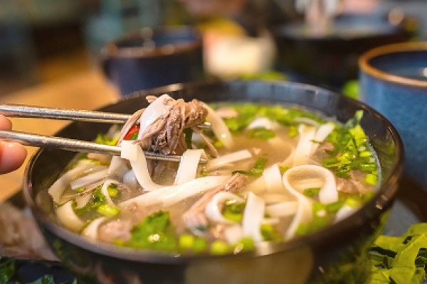 Bowl of Vietnamese pho with noodles, herbs, and beef held by chopsticks over broth.