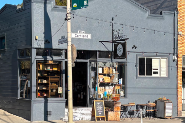 Corner café with outdoor seating, shelves of goods by the entrance, and a clock sign above the door.