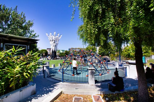 A playground with a dome climber, trees, people relaxing, and a white sculpture in the background.