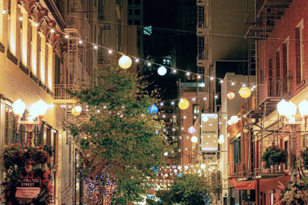 A lively city street at night with trees, string lights, and hanging lanterns decorating the scene.