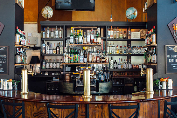 A curved wooden bar with empty chairs faces shelves stocked with bottles in a stylish, modern bar.