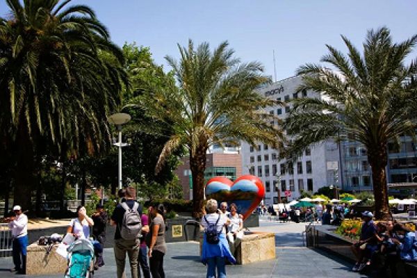 People gather near a large heart sculpture surrounded by palm trees in a sunny urban plaza.