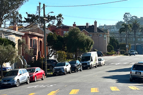 A sunny residential street with parked cars, houses, palm trees, and yellow crosswalk lines.