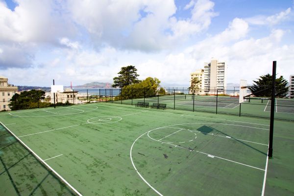 Outdoor basketball courts with faded green surfaces, surrounded by fencing, and city buildings in the background.