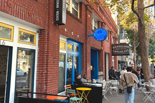 People sit at colorful tables outside a red brick café and chocolate shop on a tree-lined street.