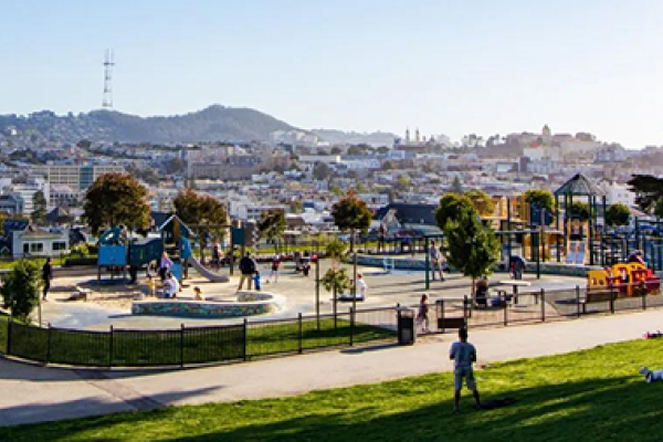 Children's playground with colorful equipment in a city park, with hills and buildings in the background.