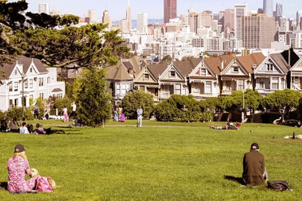 People relaxing on a grassy park with Victorian houses and a city skyline in the background.