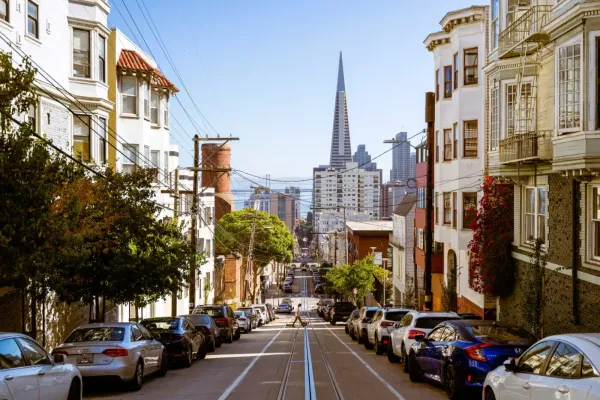 Steep San Francisco street lined with cars and buildings, with the Transamerica Pyramid in the background.