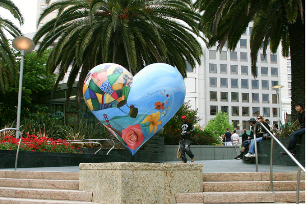 A large painted heart sculpture stands outdoors near palm trees, with people and a dog nearby.