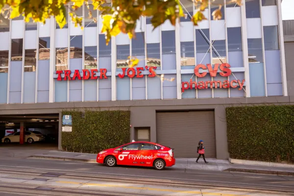 A red Flywheel car passes a Trader Joe's and CVS Pharmacy building; a person walks nearby.