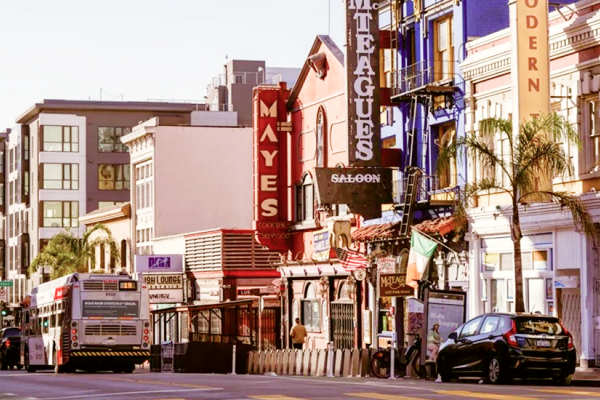 City street with colorful storefronts, a bus, parked cars, and neon signs on a sunny day.