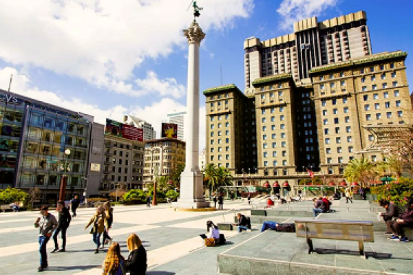 People sitting and walking in a sunny city square with tall buildings and a column in the background.