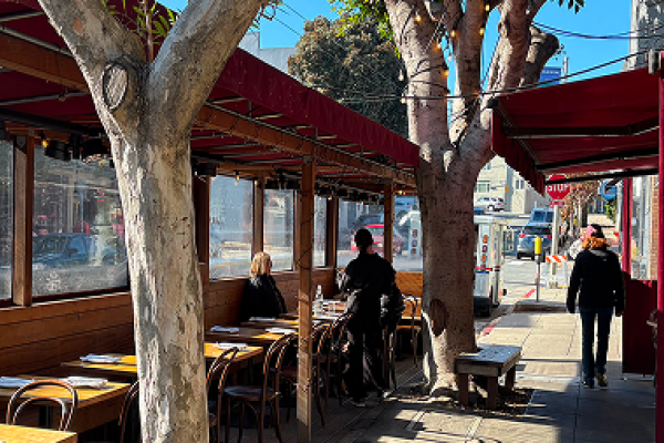 Outdoor restaurant seating with people dining under trees, next to a sidewalk on a sunny day.