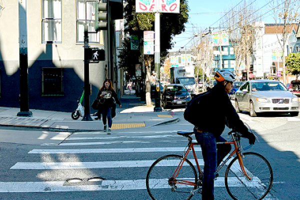 A cyclist crosses a city street at a crosswalk as pedestrians and cars wait at the intersection.