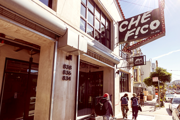 People walk past the Che Fico restaurant on a sunny street with cars parked along the sidewalk.