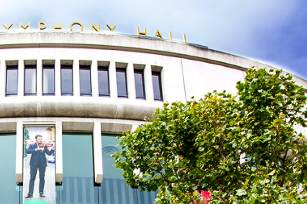 Exterior of Symphony Hall with large windows, banners, and trees in the foreground under a blue sky.