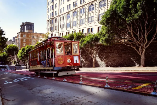 A red San Francisco cable car travels up a city street lined with trees and tall buildings.