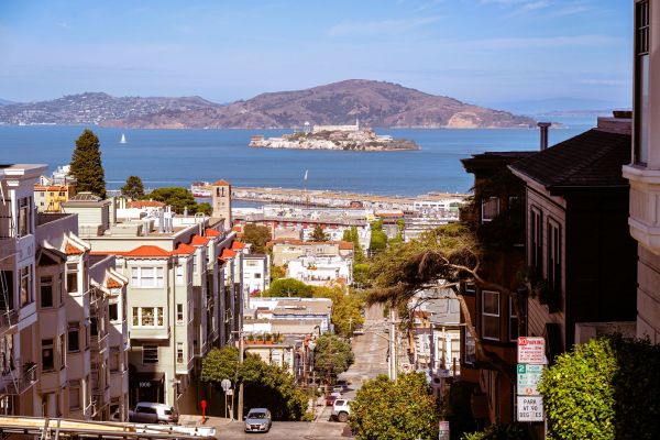 A steep San Francisco street with cars, houses, and Alcatraz Island visible in the bay under a blue sky.