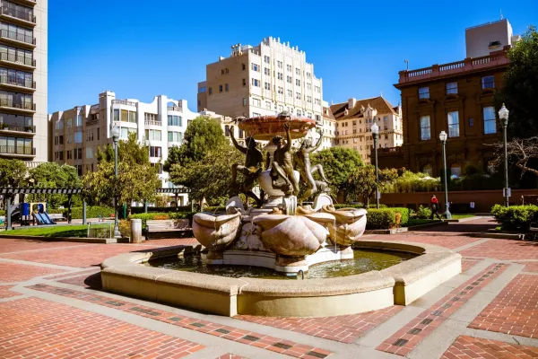 A decorative fountain with sculptures in a city park, surrounded by buildings and trees on a sunny day.