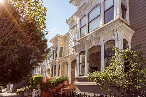 Sunlit Victorian-style houses with ornate windows and trees lining the street.