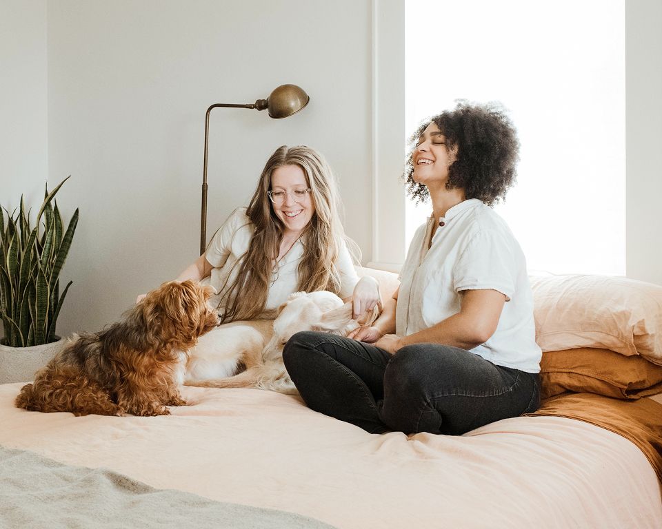 Two women smiling and sitting on a bed with two dogs in a bright, cozy bedroom with plants.