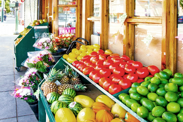 Fresh produce, including tomatoes, avocados, pineapples, and melons, displayed outside a market with flowers.