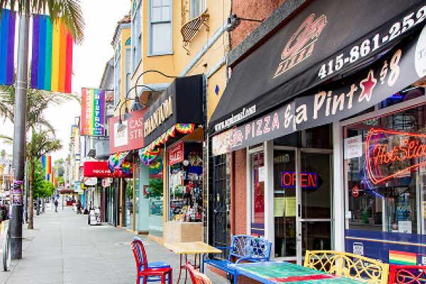 Colorful storefronts and rainbow flags line a lively street with outdoor seating and palm trees.