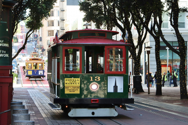 San Francisco cable cars travel down a city street lined with trees and buildings during the day.