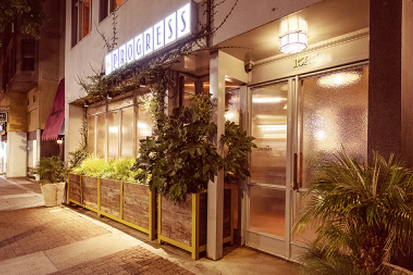Restaurant exterior at night with "PROGRESS" sign, plants in front, and warm lighting on a city sidewalk.