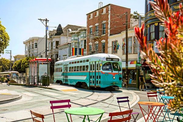 A vintage streetcar passes colorful chairs and tables on a sunny street lined with rainbow flags and buildings.
