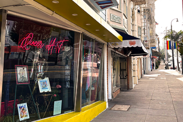 A storefront with a neon "Queer Art" sign and framed art, along a city sidewalk with rainbow flags.