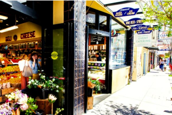 A sunny sidewalk outside a busy deli and grocery store with fresh produce and flowers on display.