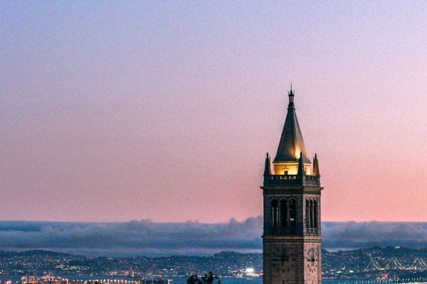 A tall clock tower with a pointed roof stands against a pastel sunset sky above a distant cityscape.