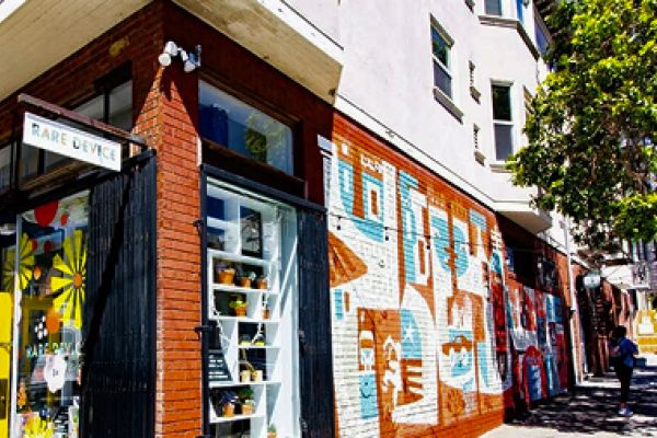 Brick building with colorful murals and a shop window, trees and people walking on a sunny sidewalk.
