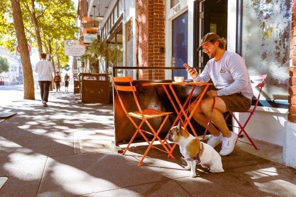 Man sitting at an outdoor café table with coffee, looking at his phone, dog sitting beside him on the sidewalk.