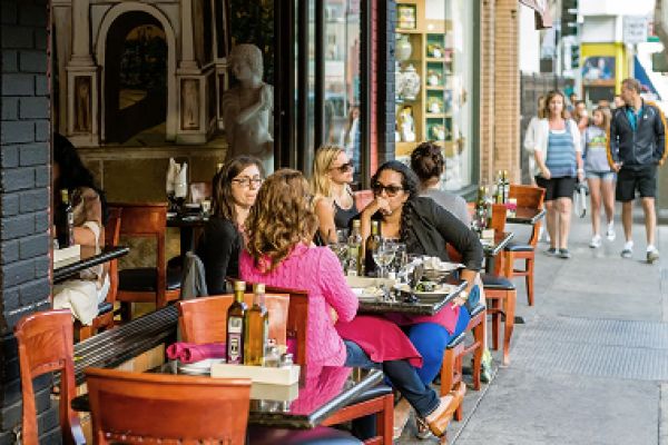 People dining at outdoor tables on a city sidewalk, with pedestrians walking by in the background.