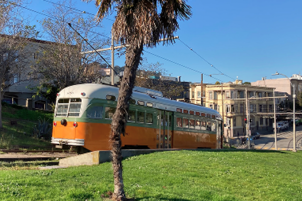 An orange and green vintage streetcar passes by a grassy park with a palm tree under a clear blue sky.
