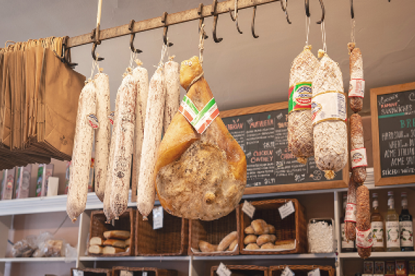 Cured meats and salamis hanging on hooks in a deli with bread and groceries in the background.