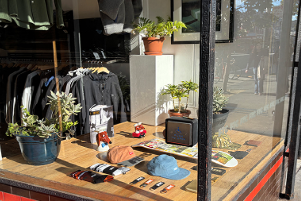 Storefront window display with hats, socks, plants, clothing on hangers, and sunlight reflecting on the glass.
