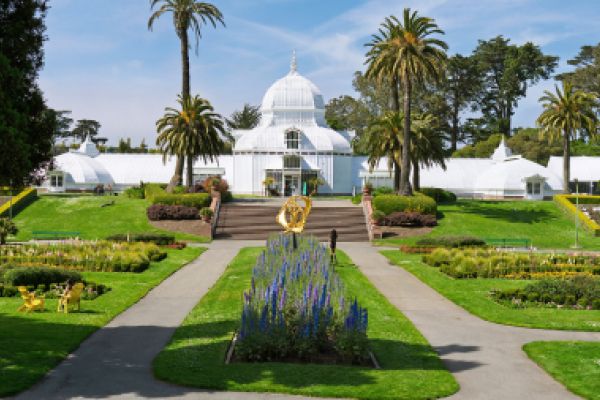A white conservatory with a glass dome, tropical plants, and gardens in front under a clear blue sky.