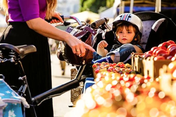 A small child in a bike seat wearing a helmet sits near fruit stands at an outdoor market with an adult.
