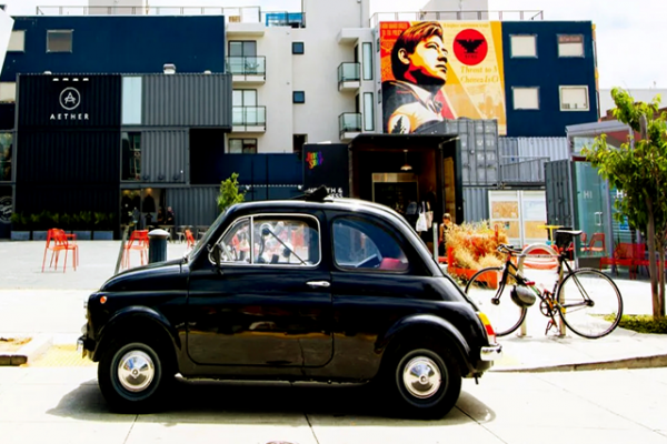 A black vintage car is parked by a bicycle near modern buildings with colorful murals in the background.