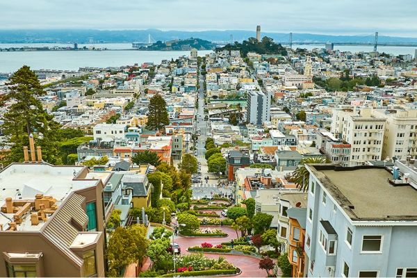 View of Lombard Street's curved section in San Francisco with buildings, trees, and the bay in the background.